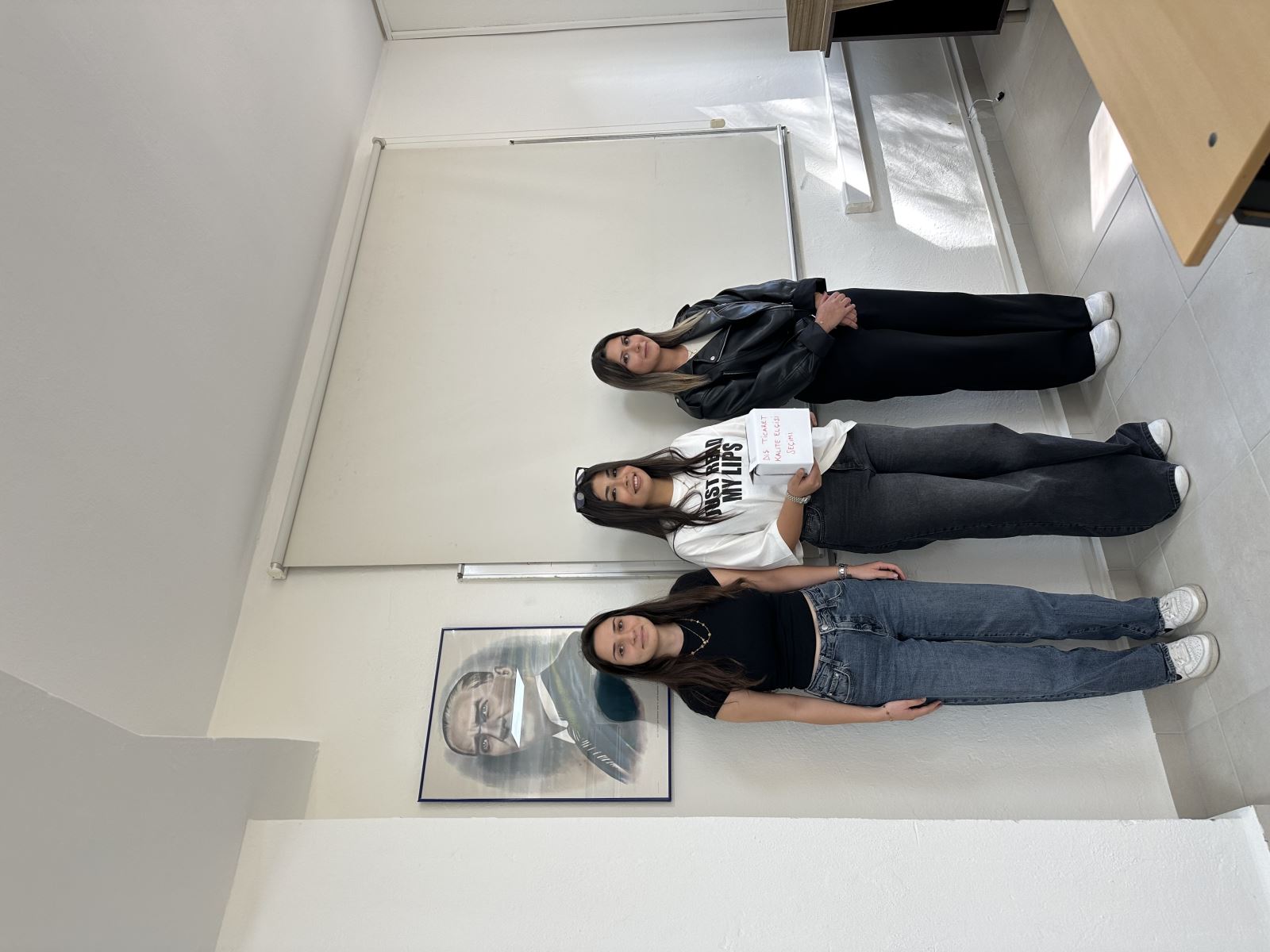 Three young women are standing side by side in a bright and simple classroom. On the wall to the left, there is a portrait of Mustafa Kemal Atatürk. In the center, a white screen or board is visible. The woman in the middle is holding a white box with the words “Foreign Trade Quality Ambassador Election” written on it. The woman on the left is wearing a black t-shirt and jeans, the one in the middle is wearing a white t-shirt and dark wide-leg jeans, and the woman on the right is dressed in a black leather jacket, black pants, and white sneakers. All three are smiling and looking at the camera.
